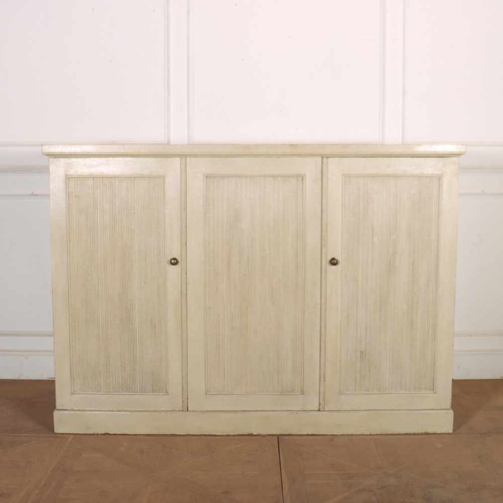 A Swedish style off-white painted wooden linen cupboard featuring three reeded panel doors with small brass knobs.