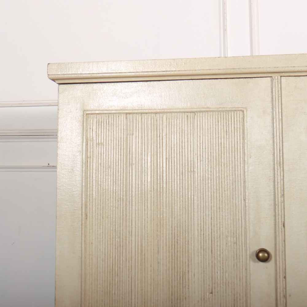 A Swedish style off-white painted wooden linen cupboard featuring three reeded panel doors with small brass knobs. View III