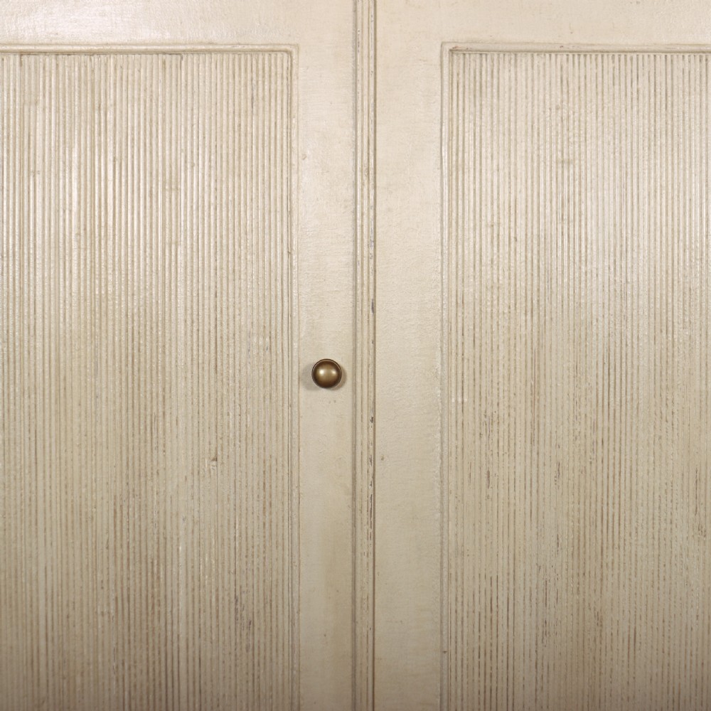 A Swedish style off-white painted wooden linen cupboard featuring three reeded panel doors with small brass knobs. View IV