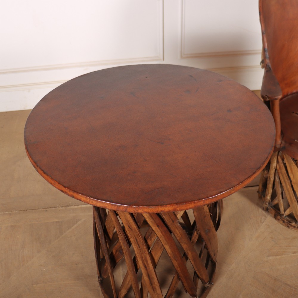A rustic furniture set featuring a curved tub armchair and a matching round side table, both constructed with diagonally woven willow slat bases and tan leather tops. View V
