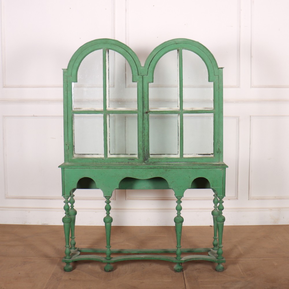 A 19th-century green painted display cabinet featuring twin arched glazed doors, a scalloped apron, and six turned legs joined by a shaped stretcher.