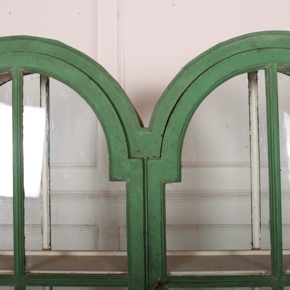 A 19th-century green painted display cabinet featuring twin arched glazed doors, a scalloped apron, and six turned legs joined by a shaped stretcher. View III