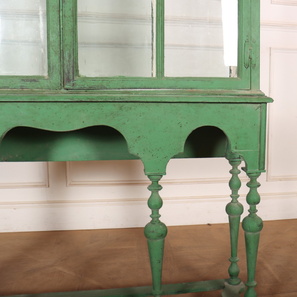 A 19th-century green painted display cabinet featuring twin arched glazed doors, a scalloped apron, and six turned legs joined by a shaped stretcher. View V