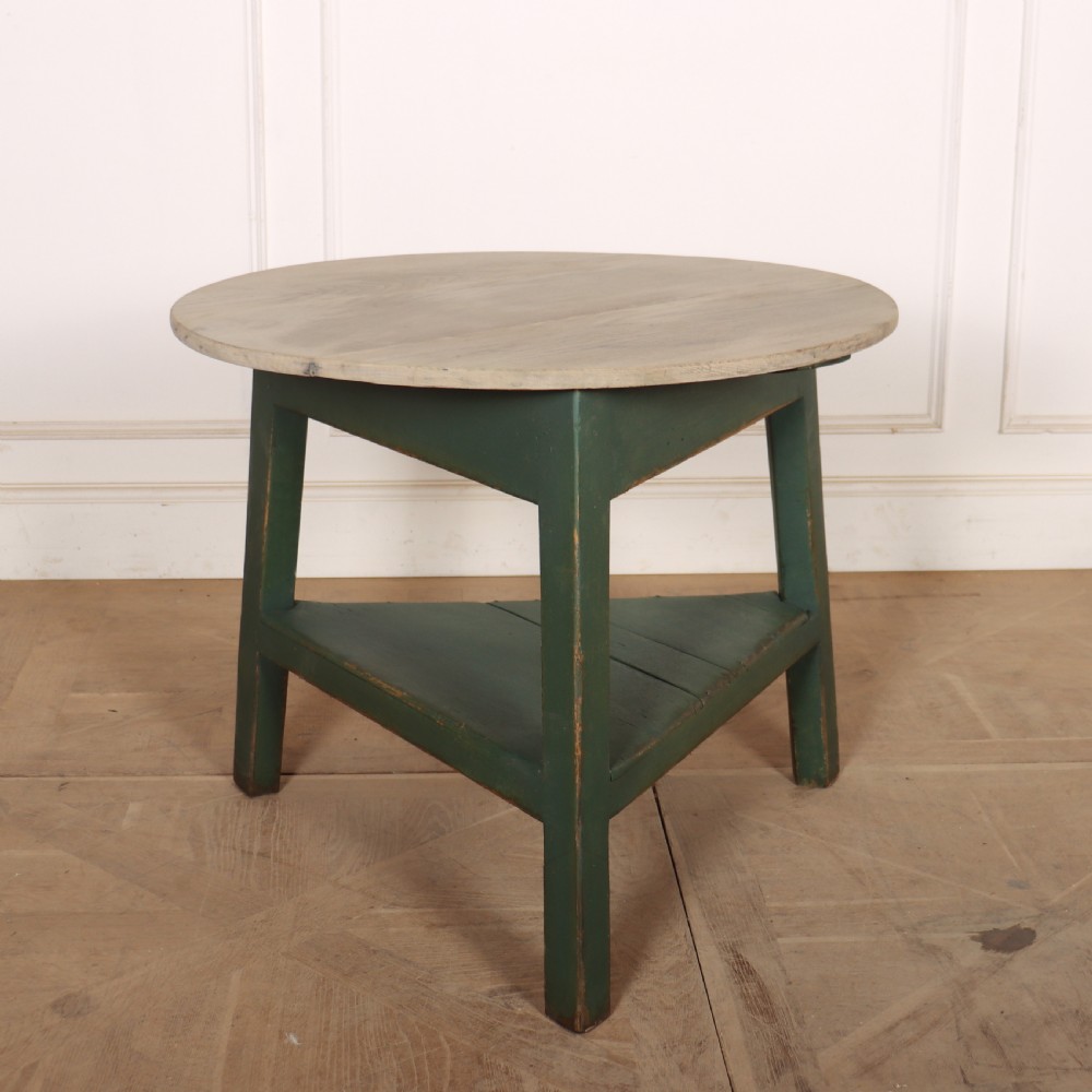A circular wooden cricket table featuring an unpainted pale timber top mounted on a dark green painted three-legged base with a lower triangular shelf.
