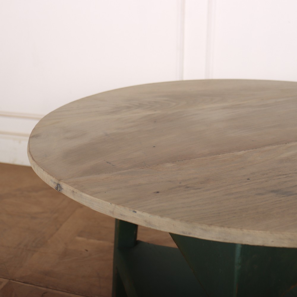 A circular wooden cricket table featuring an unpainted pale timber top mounted on a dark green painted three-legged base with a lower triangular shelf. View VI