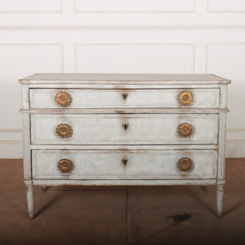 A Louis XVI style pale grey painted oak commode featuring three drawers with gilded rosette handles and fluted legs.