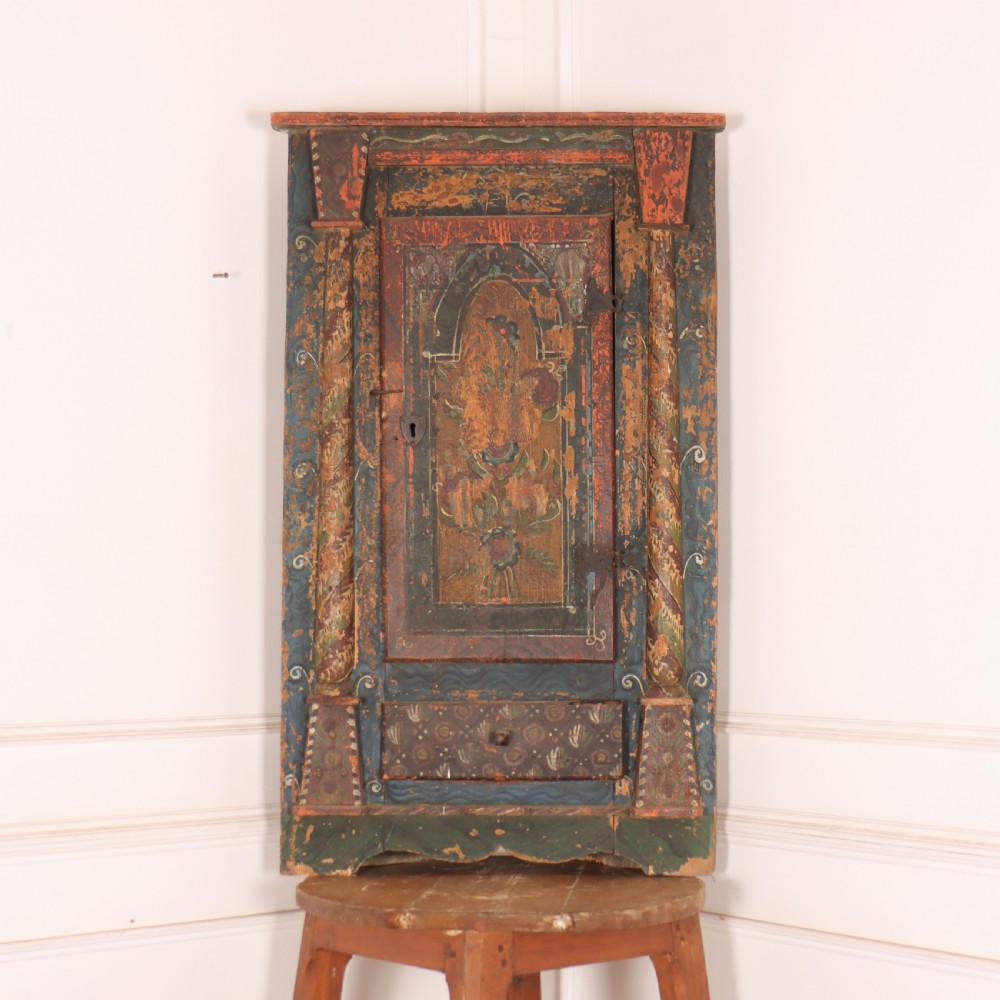An antique hand-painted Austrian pine wall cupboard featuring polychrome floral panels, carved side columns, a locking single door, and a lower drawer.