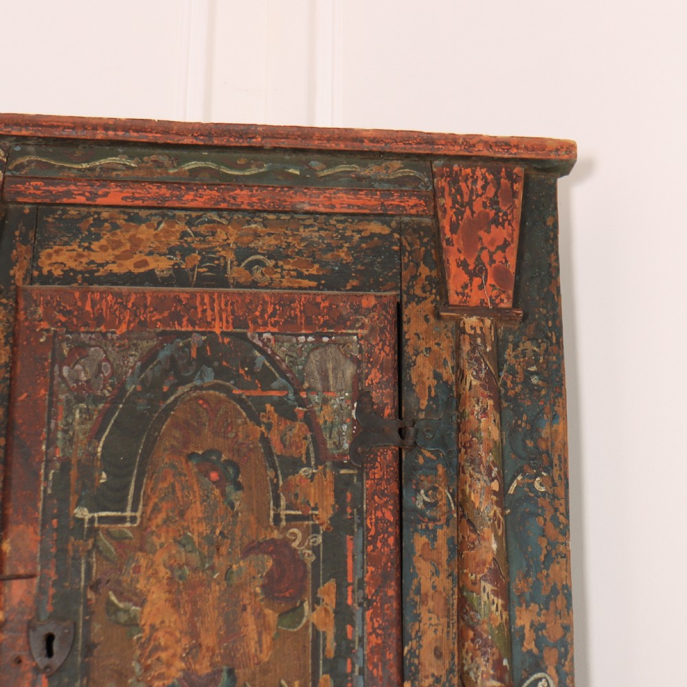 An antique hand-painted Austrian pine wall cupboard featuring polychrome floral panels, carved side columns, a locking single door, and a lower drawer. View II