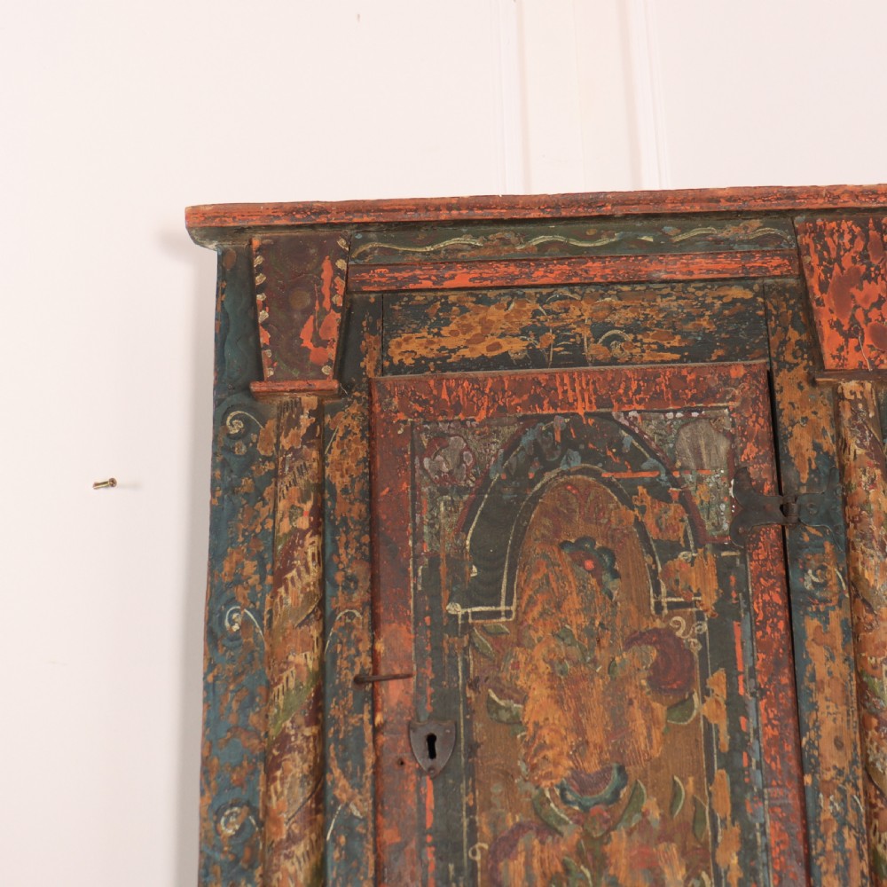 An antique hand-painted Austrian pine wall cupboard featuring polychrome floral panels, carved side columns, a locking single door, and a lower drawer. View III