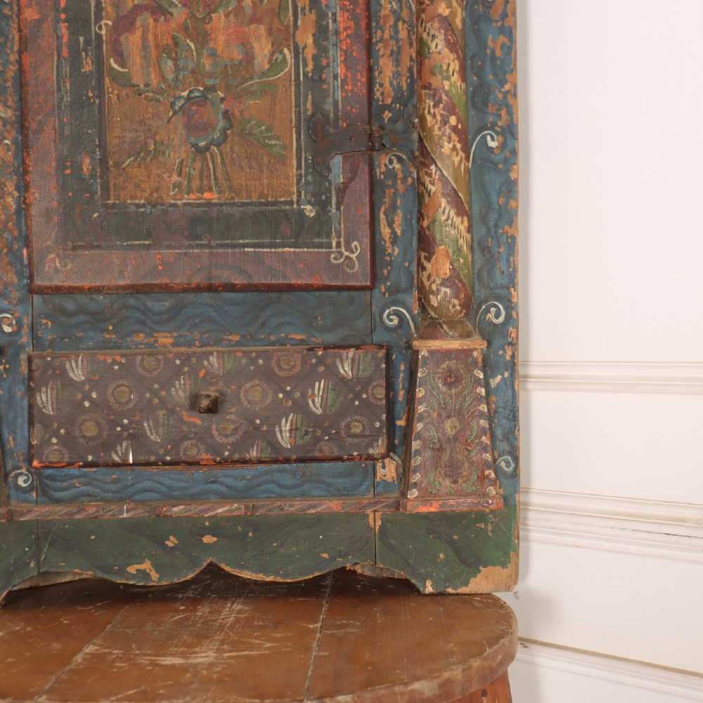 An antique hand-painted Austrian pine wall cupboard featuring polychrome floral panels, carved side columns, a locking single door, and a lower drawer. View IV