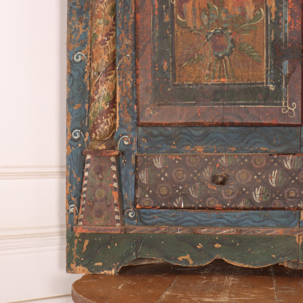 An antique hand-painted Austrian pine wall cupboard featuring polychrome floral panels, carved side columns, a locking single door, and a lower drawer. View V