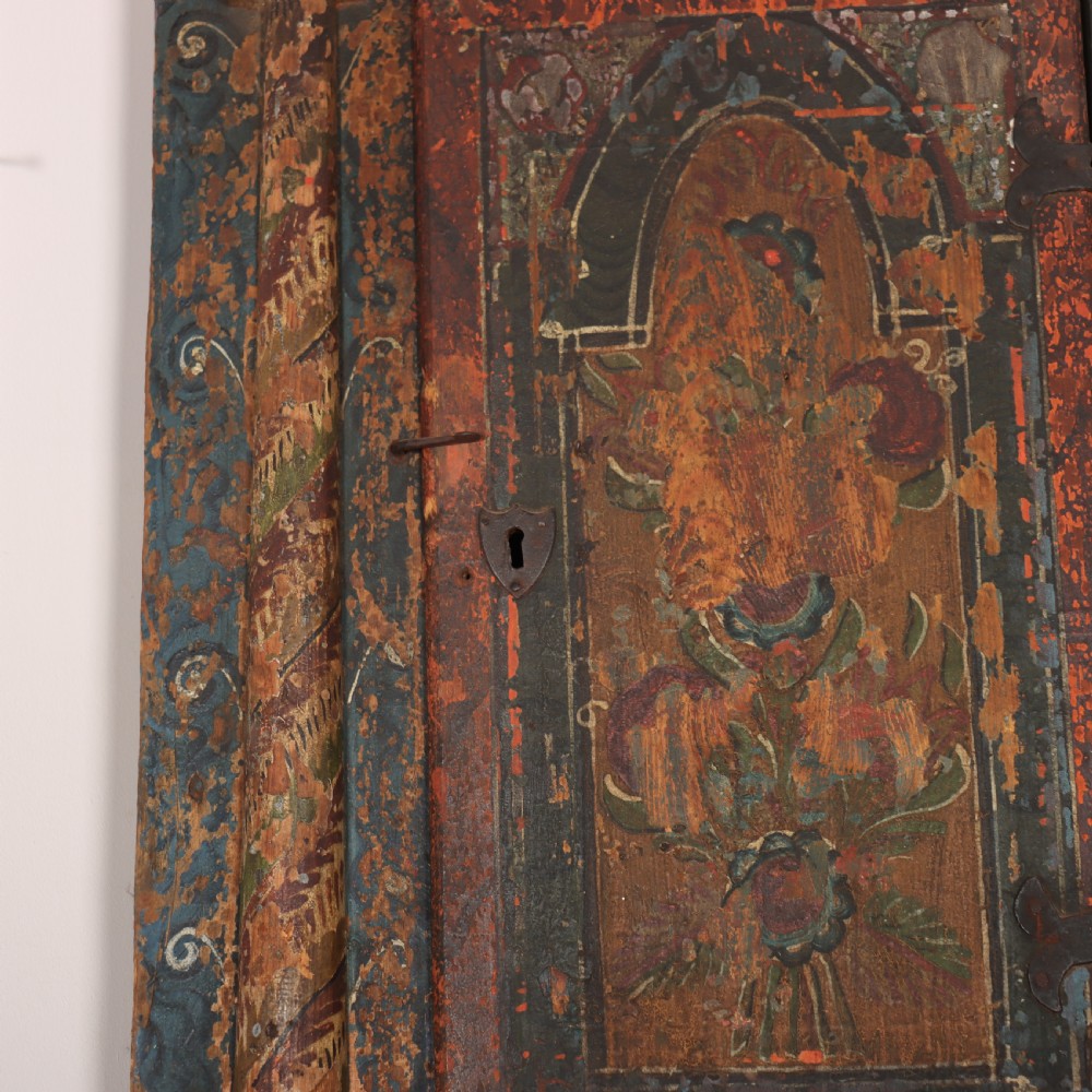 An antique hand-painted Austrian pine wall cupboard featuring polychrome floral panels, carved side columns, a locking single door, and a lower drawer. View VI