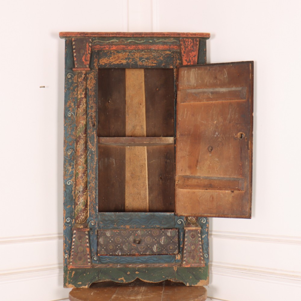 An antique hand-painted Austrian pine wall cupboard featuring polychrome floral panels, carved side columns, a locking single door, and a lower drawer. View VII