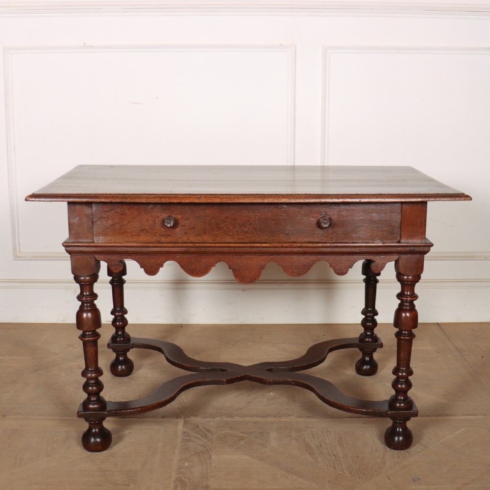 An 18th-century oak lamp table featuring a single drawer with brass knobs, a scalloped apron, turned legs, and an X-frame cross stretcher.
