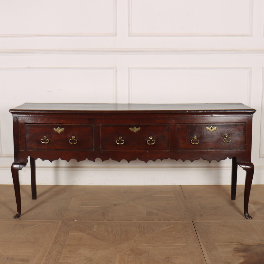 An 18th-century dark oak dresser base featuring three drawers, brass drop handles, keyhole escutcheons, a scalloped lower apron, and cabriole legs.