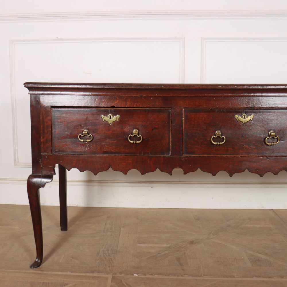 An 18th-century dark oak dresser base featuring three drawers, brass drop handles, keyhole escutcheons, a scalloped lower apron, and cabriole legs. View II
