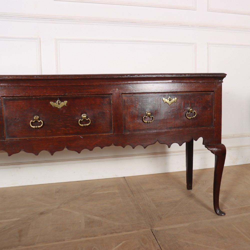 An 18th-century dark oak dresser base featuring three drawers, brass drop handles, keyhole escutcheons, a scalloped lower apron, and cabriole legs. View IV
