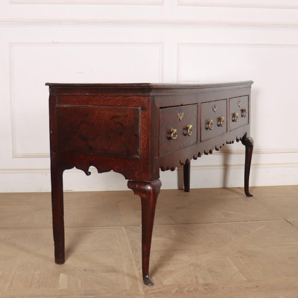 An 18th-century dark oak dresser base featuring three drawers, brass drop handles, keyhole escutcheons, a scalloped lower apron, and cabriole legs. View VI