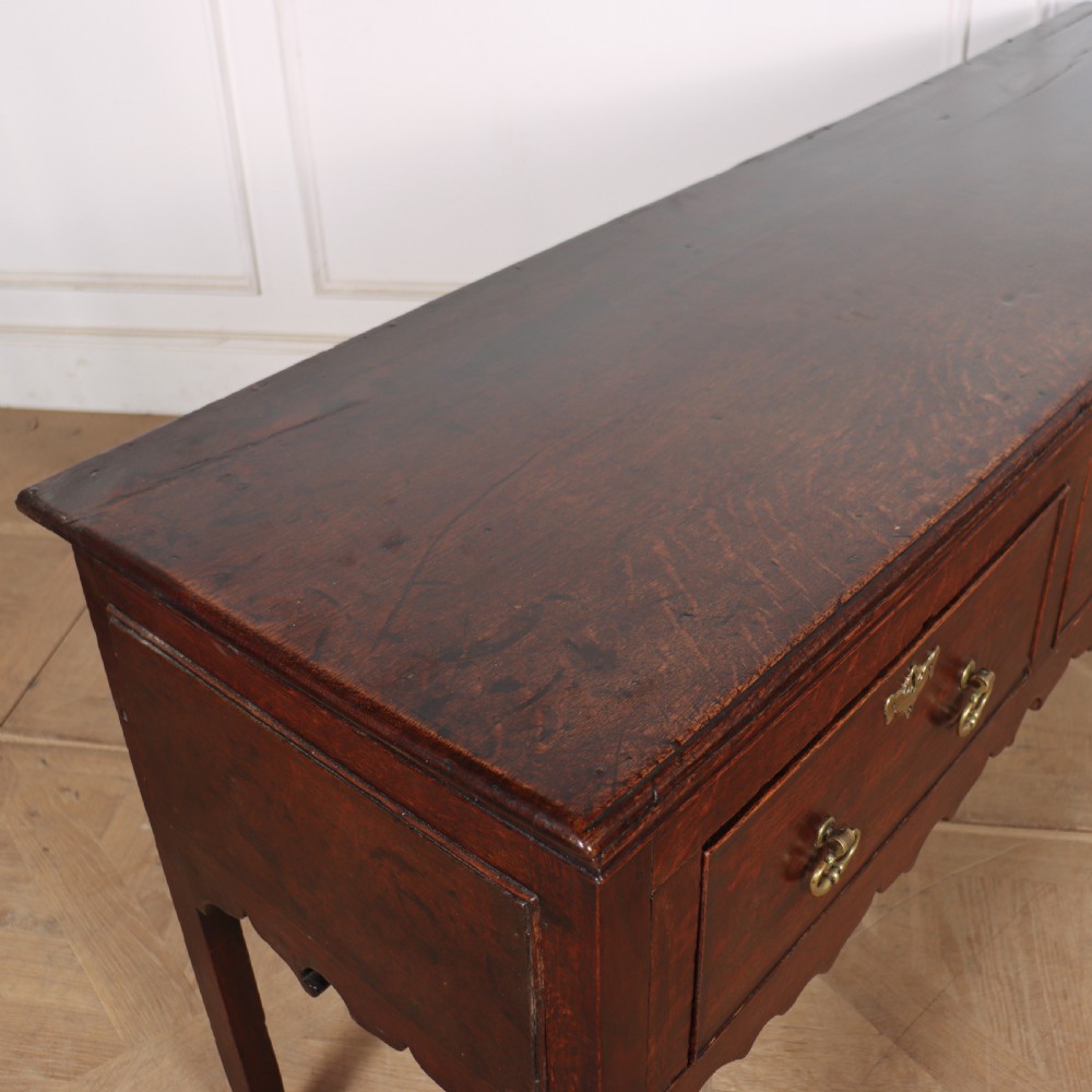 An 18th-century dark oak dresser base featuring three drawers, brass drop handles, keyhole escutcheons, a scalloped lower apron, and cabriole legs. View VIII