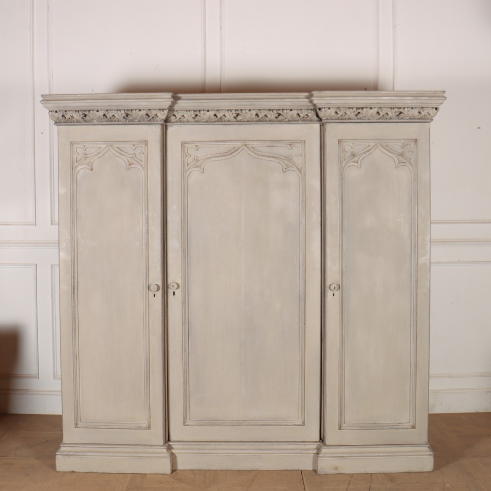 A large 19th-century painted wooden three-door wardrobe featuring carved Gothic arches and a detailed crown moulding cornice.