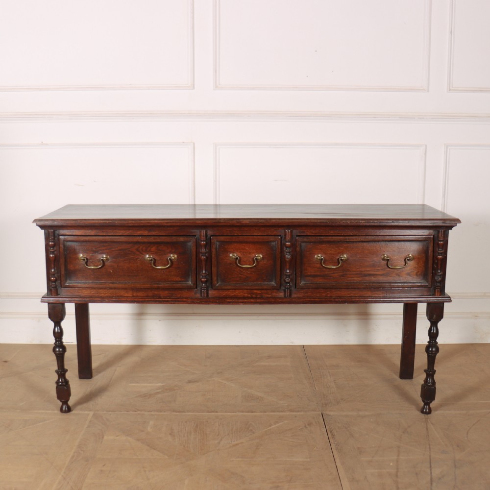 A dark oak dresser base featuring three panelled drawers with brass handles and turned front legs.