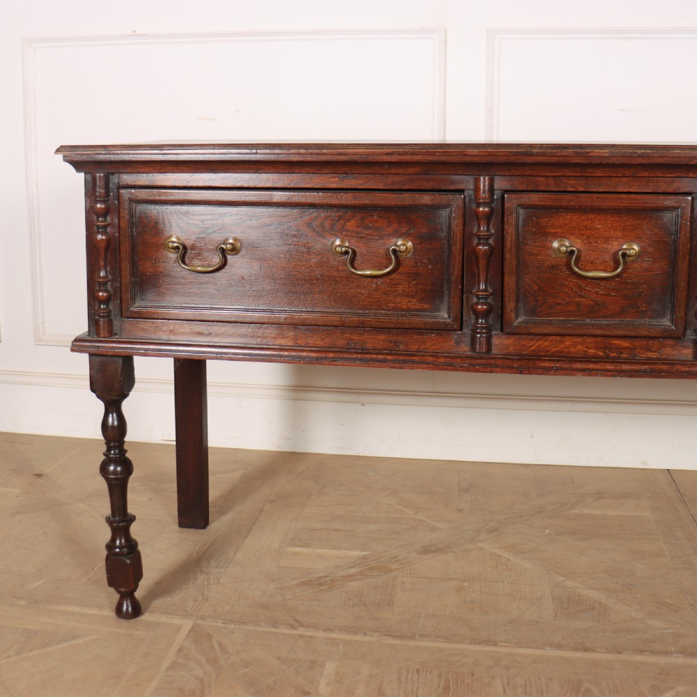 A dark oak dresser base featuring three panelled drawers with brass handles and turned front legs. View II