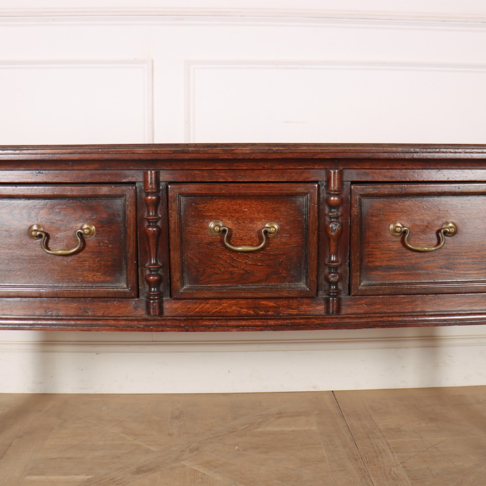 A dark oak dresser base featuring three panelled drawers with brass handles and turned front legs. View III