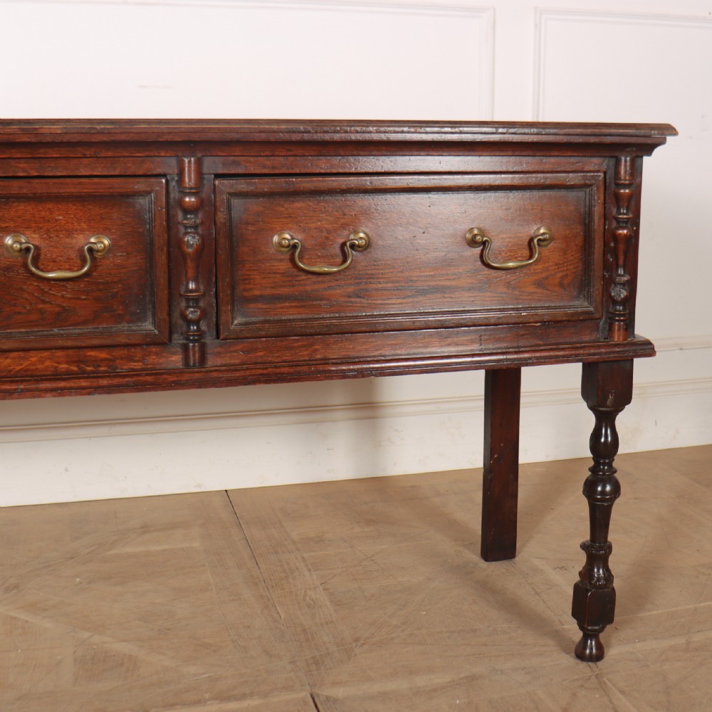 A dark oak dresser base featuring three panelled drawers with brass handles and turned front legs. View IV