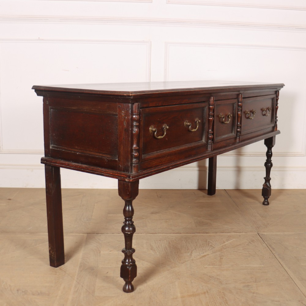 A dark oak dresser base featuring three panelled drawers with brass handles and turned front legs. View V