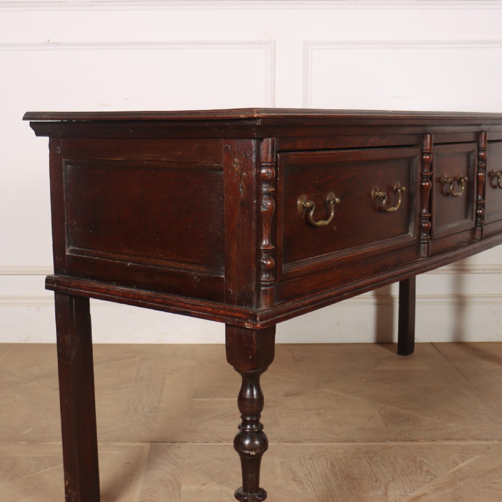 A dark oak dresser base featuring three panelled drawers with brass handles and turned front legs. View VI