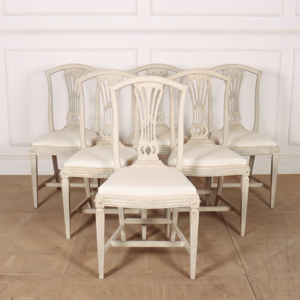 A set of six Gustavian style dining chairs painted in distressed off-white with carved splat backs and white upholstered seats.