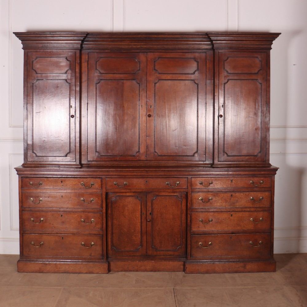 An antique English oak housekeepers cupboard featuring a breakfront upper section with fielded panel doors above a base with multiple drawers and brass hardware.