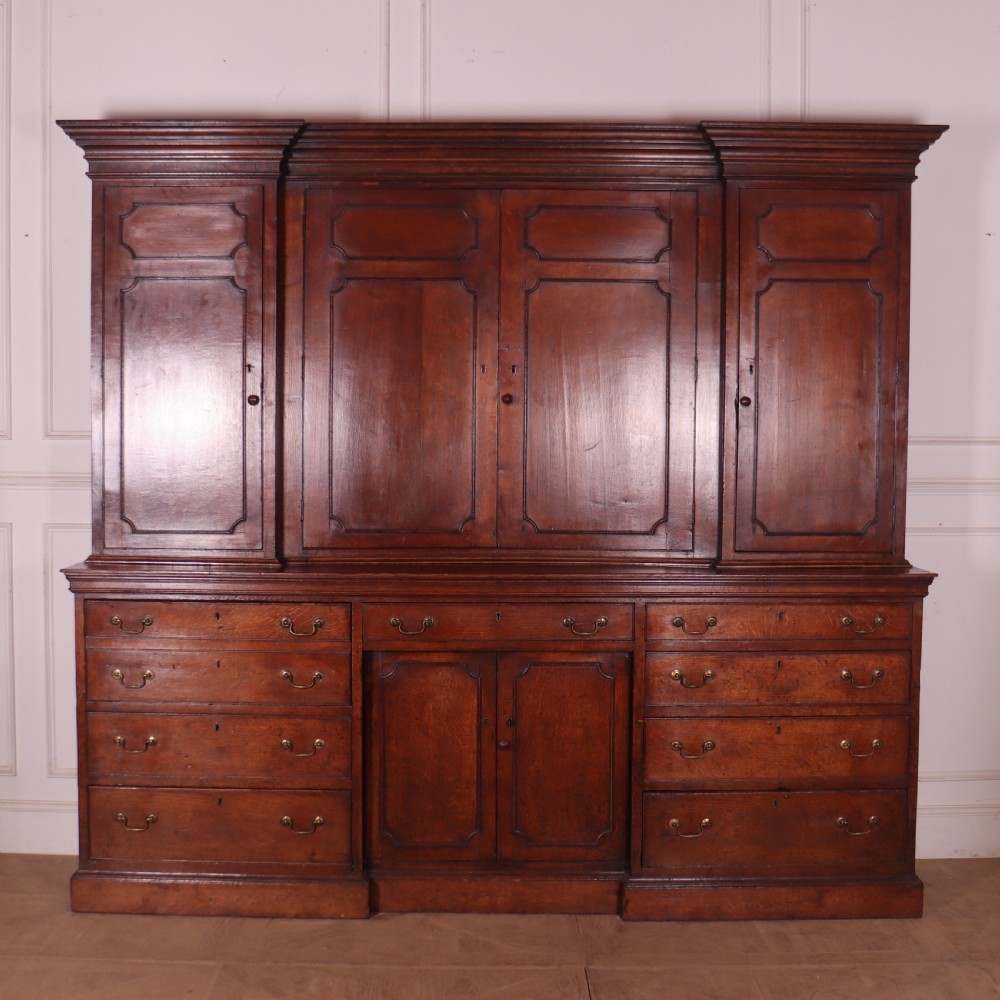 An antique English oak housekeepers cupboard featuring a breakfront upper section with fielded panel doors above a base with multiple drawers and brass hardware. View XV
