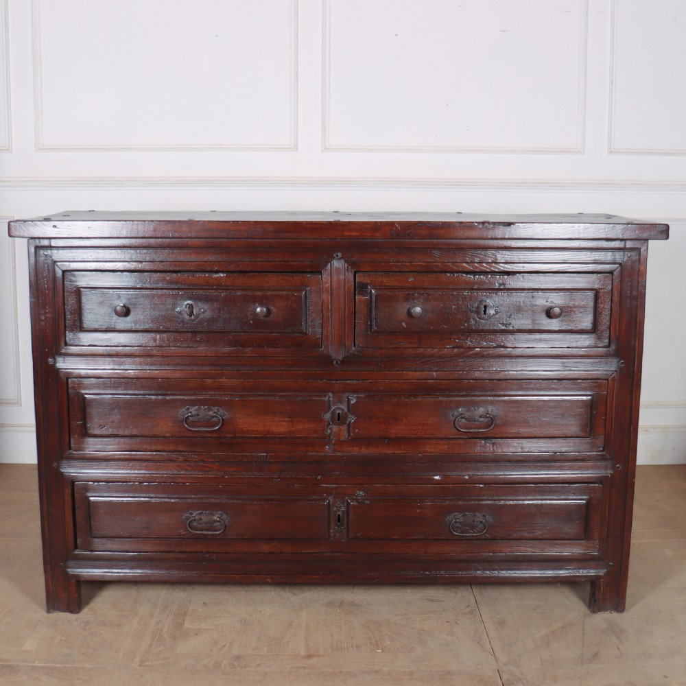 A large dark brown Spanish oak commode featuring deep geometric drawer mouldings, central lock escutcheons, and iron drop handles.