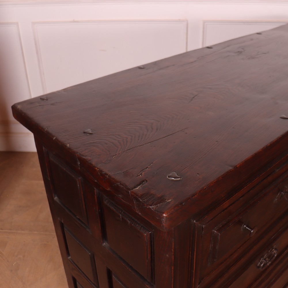 A large dark brown Spanish oak commode featuring deep geometric drawer mouldings, central lock escutcheons, and iron drop handles. View VIII