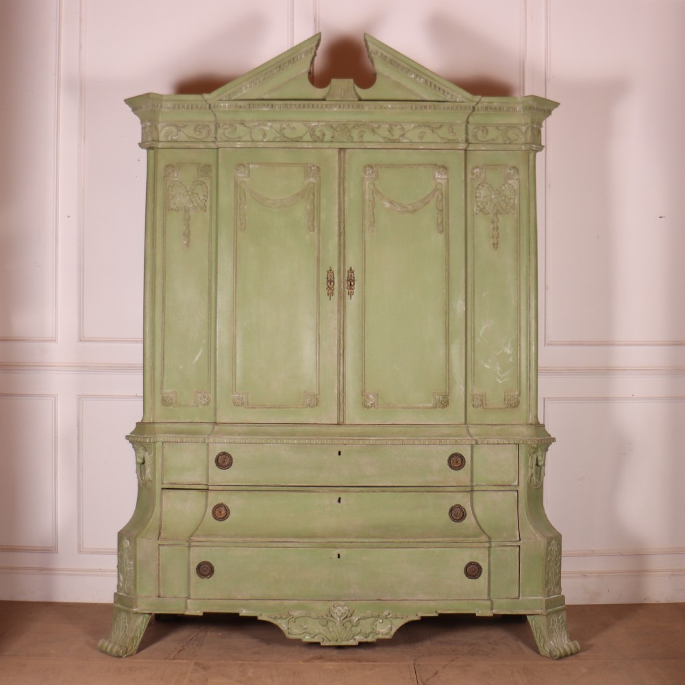 A large sage green painted Dutch linen cupboard featuring a broken pediment cornice, carved swag door panels, brass drop handles, and three lower drawers.