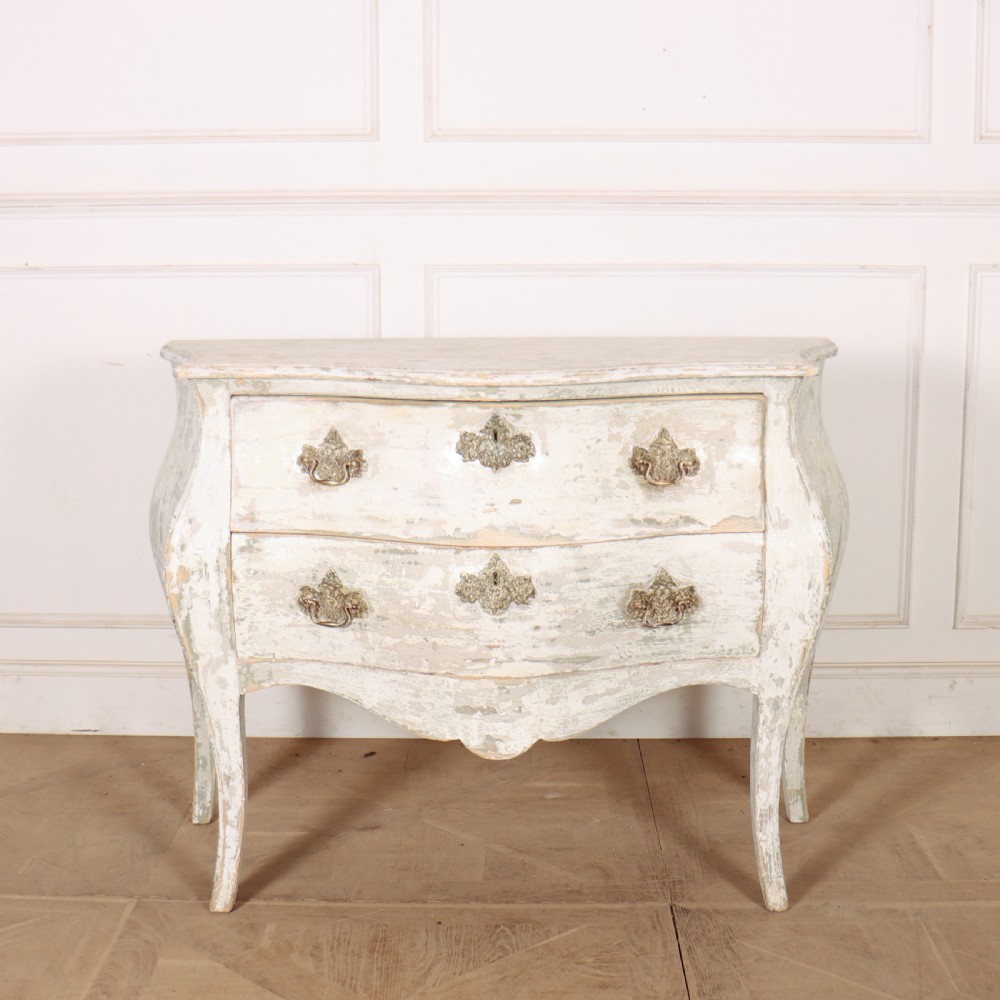 A French painted two-drawer commode featuring a bombe shape, distressed off-white finish, carved apron, cabriole legs, and ornate brass hardware.