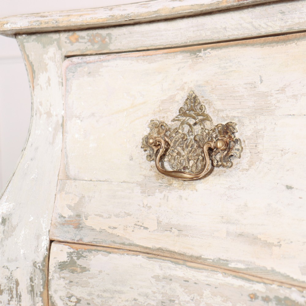 A French painted two-drawer commode featuring a bombe shape, distressed off-white finish, carved apron, cabriole legs, and ornate brass hardware. View V