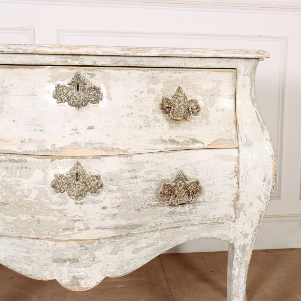 A French painted two-drawer commode featuring a bombe shape, distressed off-white finish, carved apron, cabriole legs, and ornate brass hardware. View VI