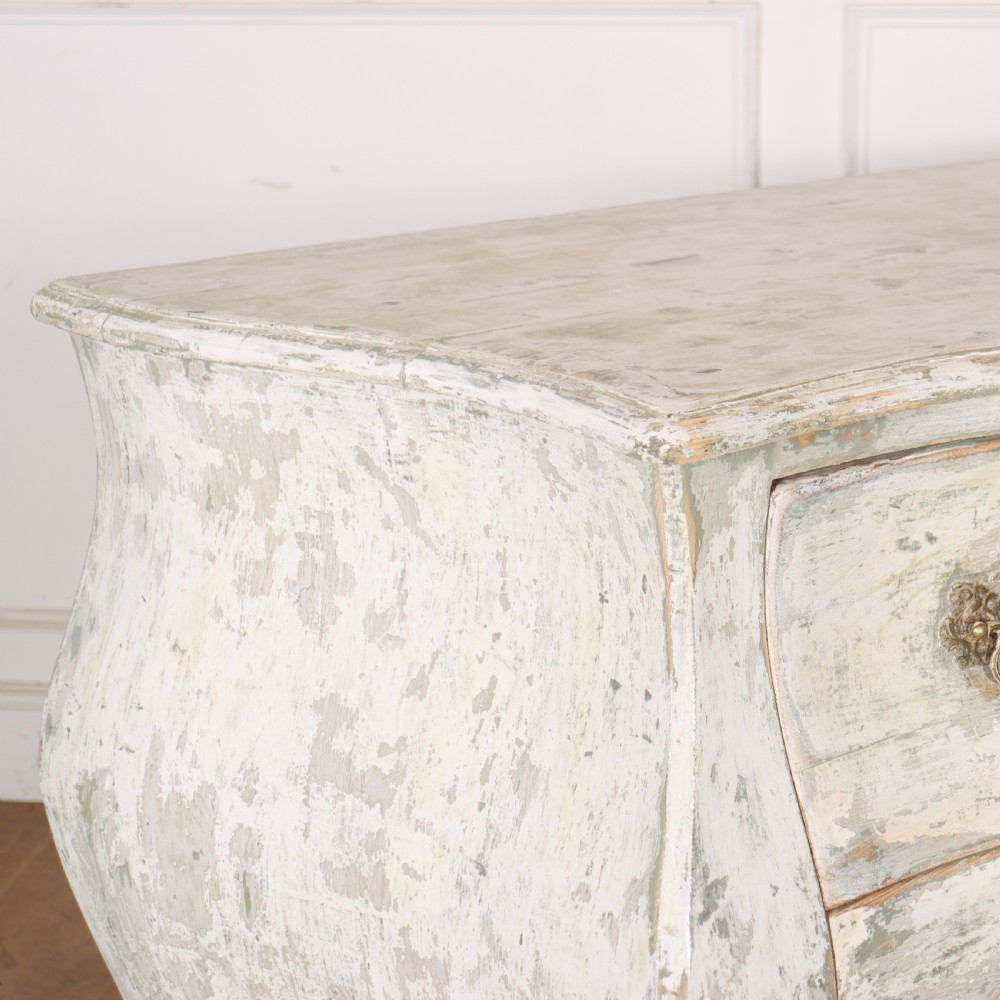 A French painted two-drawer commode featuring a bombe shape, distressed off-white finish, carved apron, cabriole legs, and ornate brass hardware. View VII