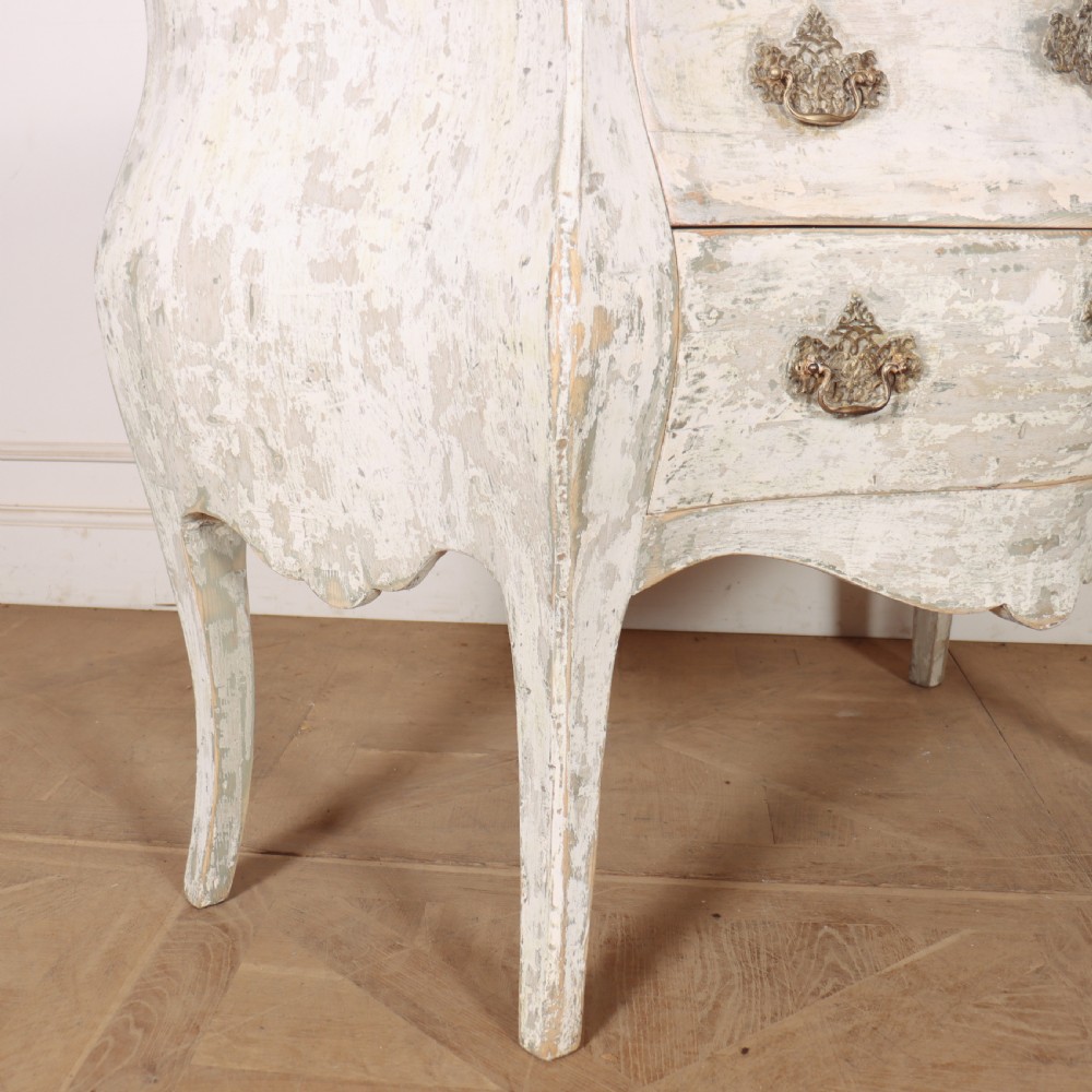 A French painted two-drawer commode featuring a bombe shape, distressed off-white finish, carved apron, cabriole legs, and ornate brass hardware. View VIII