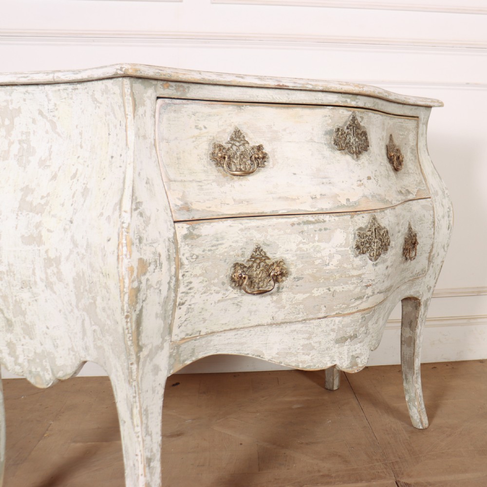 A French painted two-drawer commode featuring a bombe shape, distressed off-white finish, carved apron, cabriole legs, and ornate brass hardware. View IX