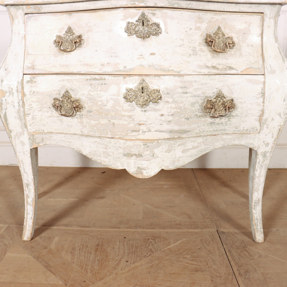 A French painted two-drawer commode featuring a bombe shape, distressed off-white finish, carved apron, cabriole legs, and ornate brass hardware. View X