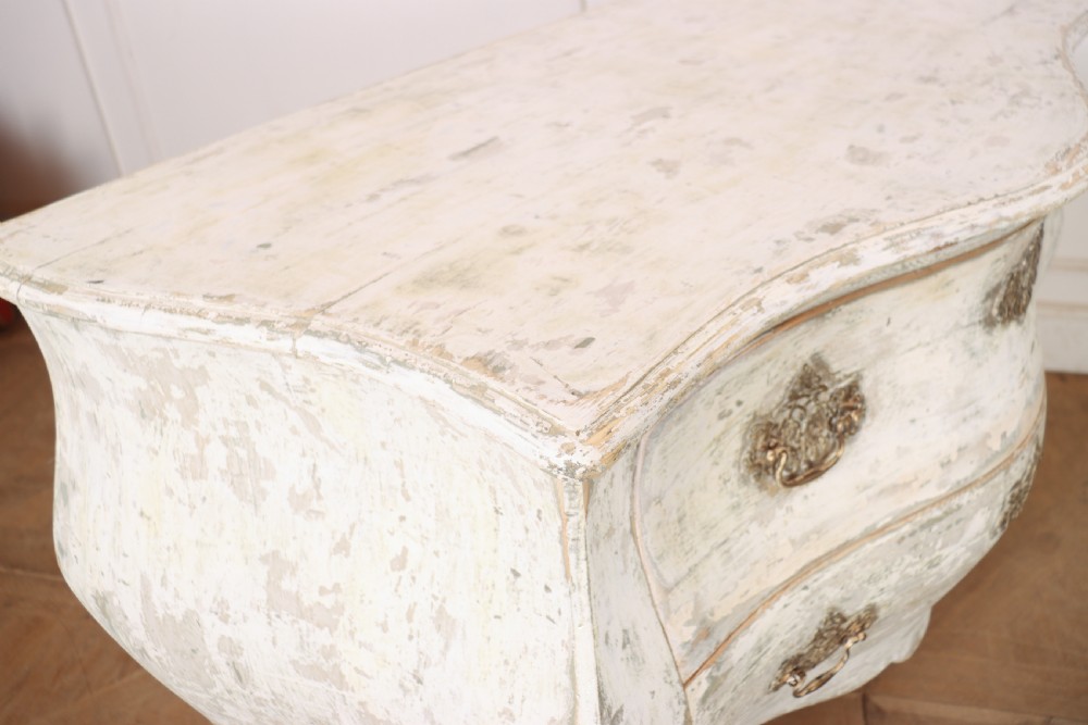 A French painted two-drawer commode featuring a bombe shape, distressed off-white finish, carved apron, cabriole legs, and ornate brass hardware. View XI