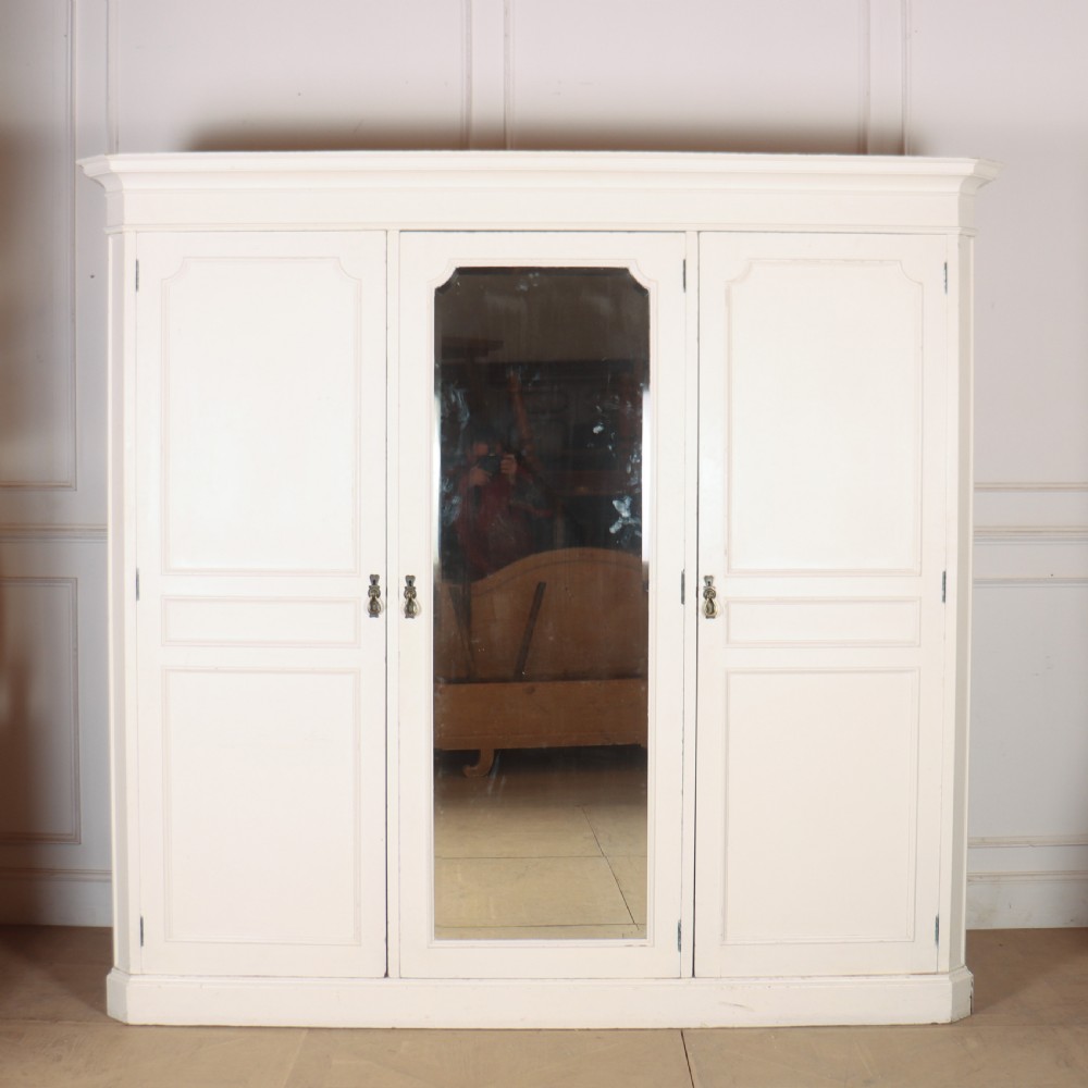 A large 19th-century off-white painted wardrobe featuring three doors, a central arched full-length mirror, moulded cornice, and drop handles.