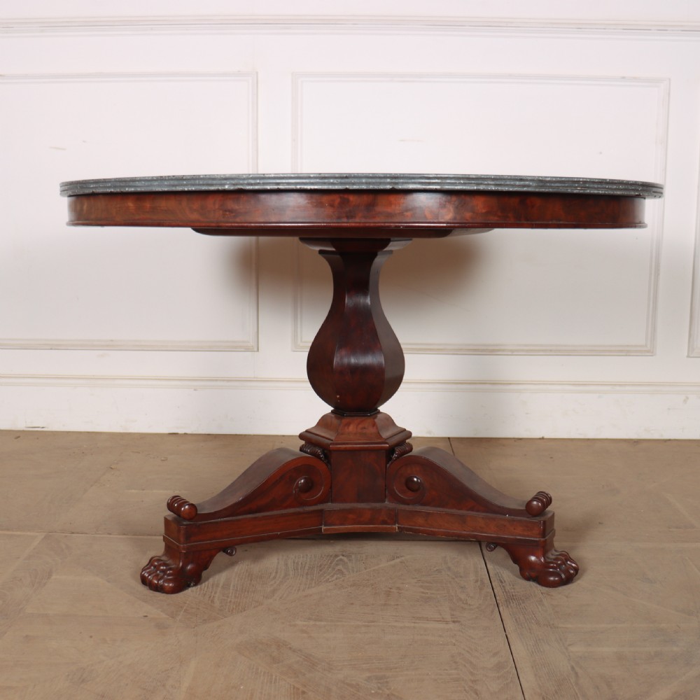 A large circular French gueridon table featuring a dark grey marble top set upon a dark mahogany pedestal base with a triform platform and carved paw feet.