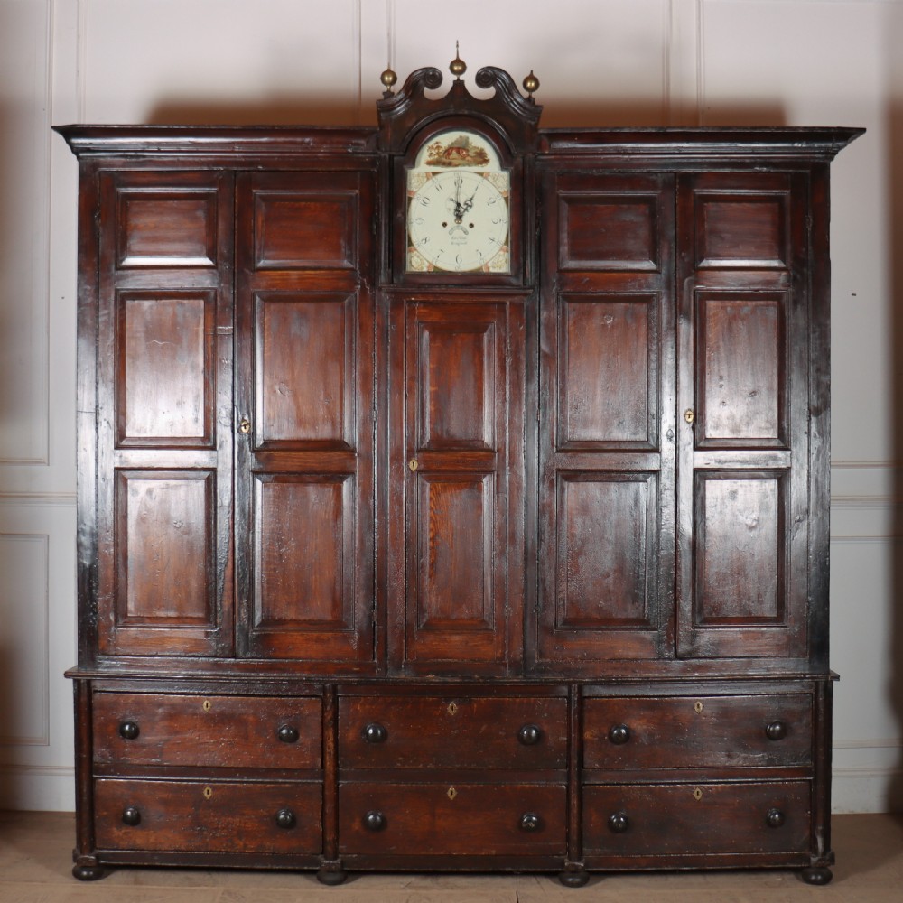 An antique dark brown wooden housekeepers cupboard featuring panelled doors, a central integrated grandfather clock, and a base section with six drawers.