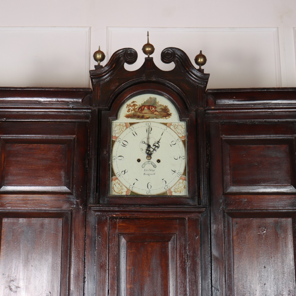 An antique dark brown wooden housekeepers cupboard featuring panelled doors, a central integrated grandfather clock, and a base section with six drawers. View III
