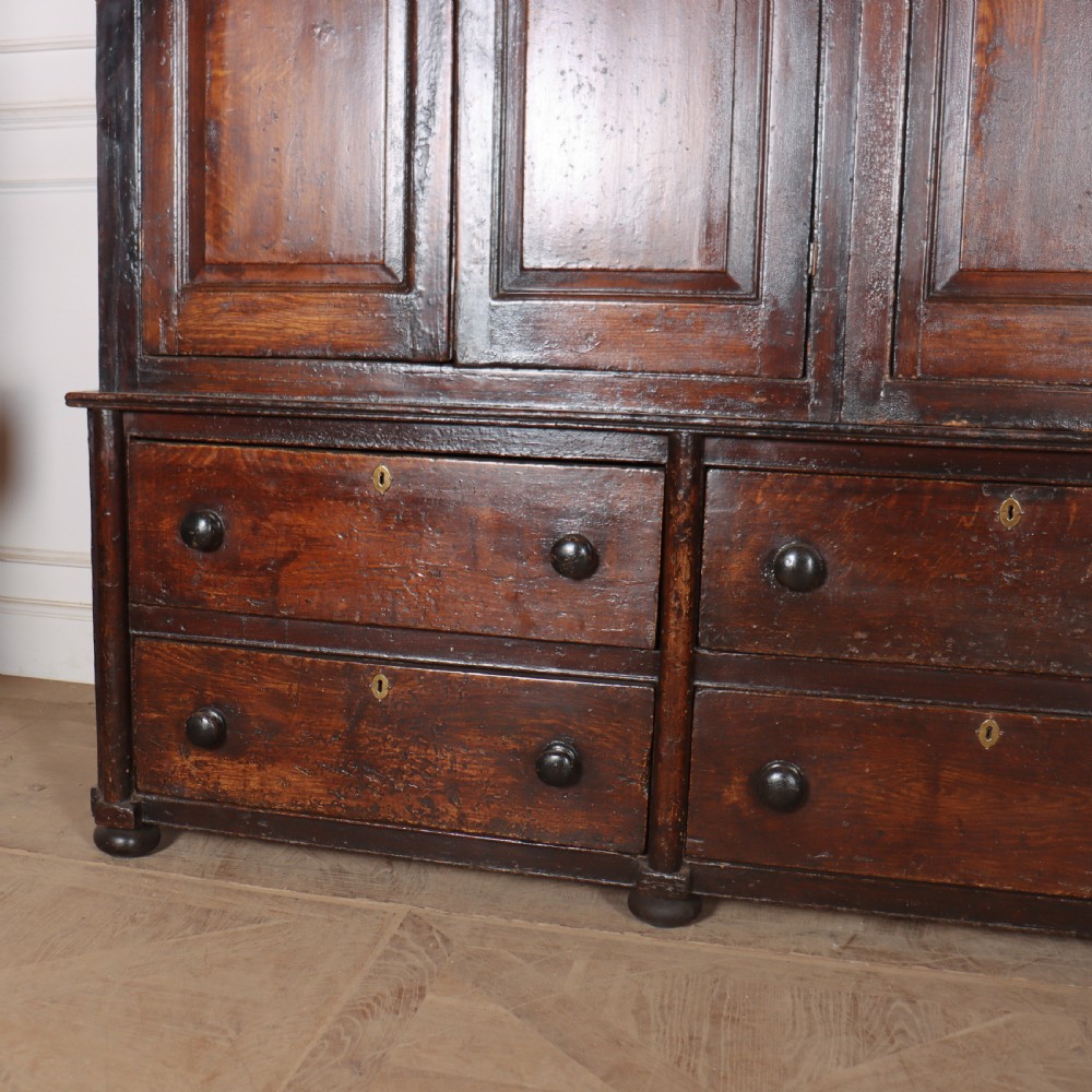 An antique dark brown wooden housekeepers cupboard featuring panelled doors, a central integrated grandfather clock, and a base section with six drawers. View V
