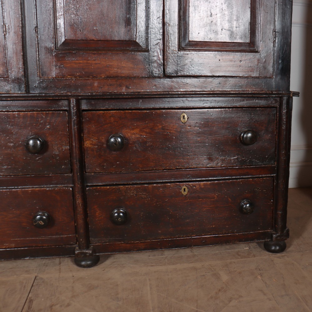An antique dark brown wooden housekeepers cupboard featuring panelled doors, a central integrated grandfather clock, and a base section with six drawers. View VI